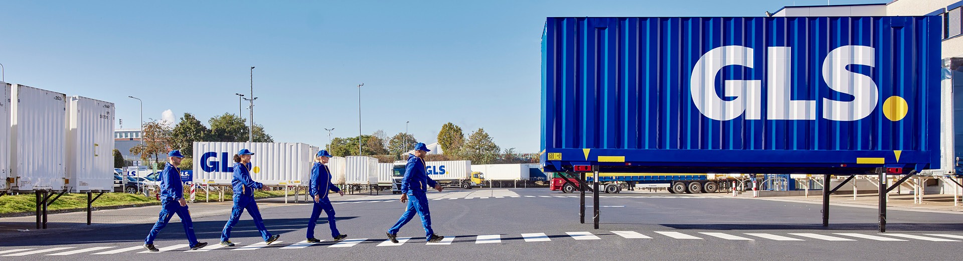 Équipe GLS sur site logistique traversant une zone sécurisée devant une semi-remorque bleue, centre de distribution et transport de colis.