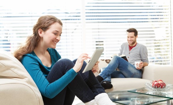 A woman using a tablet and a man reading a book while sitting on a couch.